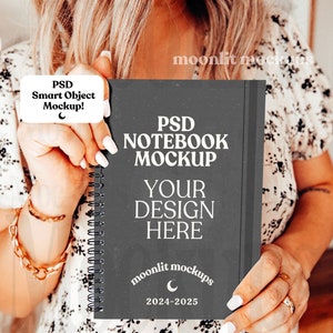 May include: A black notebook mockup with a white spiral binding. The notebook cover has the text "PSD NOTEBOOK MOCKUP" and "YOUR DESIGN HERE" in white. The bottom of the cover has a crescent moon and the text "moonlit mockups 2024-2025".
