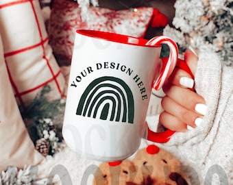 May include: White ceramic mug with a red interior and handle, featuring a green rainbow design and the text "YOUR DESIGN HERE". A candy cane is placed on the handle. The mug is held in front of a Christmas tree.