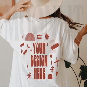 以下が含まれることがあります： 赤いグラフィックデザインが施された白いオーバーサイズTシャツ。デザインは「YOUR DESIGN HERE」と書いてあり、抽象的な形と星で囲まれています。