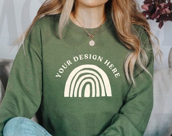 Puede incluir: Una mujer lleva una sudadera verde con un diseño de arcoíris blanco y el texto "YOUR DESIGN HERE". Está sentada en un sofá con un cojín blanco y un cojín gris.