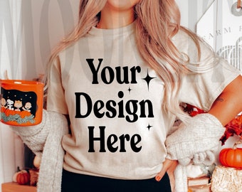 May include: A beige t-shirt with the text "Your Design Here" in black. An orange mug with a cartoon design is held in the left hand. The person is wearing black leggings and a gray cardigan. The background includes fall decorations.