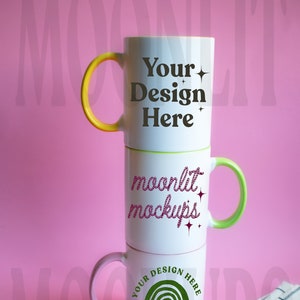 Op de afbeelding: Drie witte keramische mokken gestapeld op elkaar op een roze oppervlak. De bovenste mok heeft een gele handgreep en de tekst "Your Design Here" in zwart. De middelste mok heeft een groene handgreep en de tekst "moonlit mockups" in roze. De onderste mok heeft een roze handgreep en een groene regenboog met de tekst "Your Design Here" in zwart.