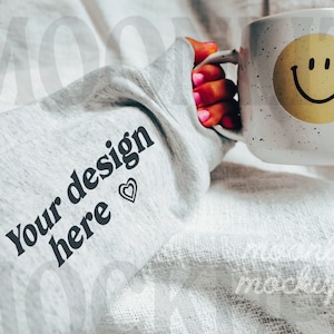 May include: A gray sweatshirt with the text "Your design here" and a small heart printed on the sleeve. The sweatshirt is folded and a hand is holding a white mug with a yellow smiley face on it.