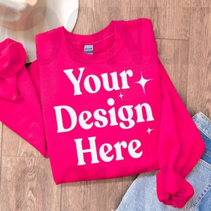 May include: A bright pink sweatshirt with the text "Your Design Here" in white. The sweatshirt is laid flat on a wood floor with a pair of blue jeans and a white hat.