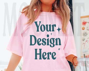 Pode incluir: Uma camiseta rosa oversized com o texto "Your Design Here" em azul escuro. A camiseta está sendo usada por uma pessoa e tem um caimento relaxado.