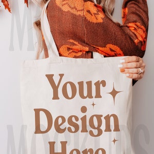 Może przedstawiać: Biała torba typu tote z płótna z brązowymi uchwytami. Torba ma napis "Your Design Here" nadrukowany z przodu brązową czcionką.