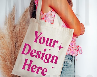 May include: A woman is holding a white canvas tote bag with pink text that says "Your Design Here". The bag has pink glitter accents. The woman is wearing a pink floral top and blue jeans.
