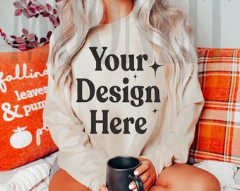 May include: A woman wearing a beige sweatshirt with the text "Your Design Here" on it. She is sitting on a couch with a blanket and holding a black mug.