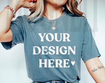 May include: A woman wearing a blue t-shirt with the text "YOUR DESIGN HERE" in white. She is wearing blue jeans and a plaid blanket is draped over her legs.