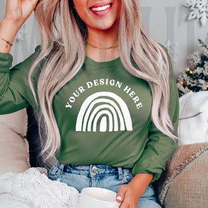 May include: A woman wearing a green long-sleeved shirt with a white rainbow design and the text "YOUR DESIGN HERE". She is sitting on a couch and holding a white mug.