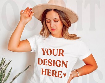 May include: A white t-shirt with the text "YOUR DESIGN HERE" in brown lettering. The shirt is being worn by a person with a brown hat and blue jeans.