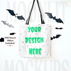 May include: White canvas tote bag with black handles hanging on a white wall. The bag has a black and white Halloween themed pattern and the text "YOUR DESIGN HERE" in green. There are black bats flying around the bag.
