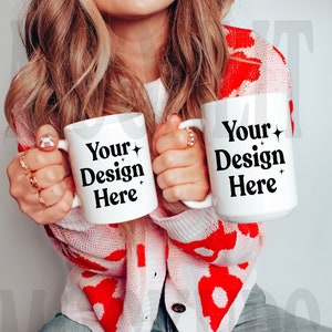 以下が含まれることがあります： 白いセラミック製のマグカップ2つ。黒い文字で「Your Design Here」と書いてあり、白い背景になっています。マグカップは、白と赤のセーターを着た人が持っています。