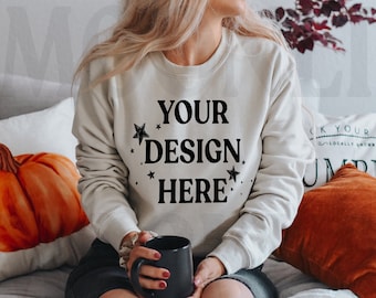 May include: A white sweatshirt with the text "YOUR DESIGN HERE" in black lettering and small stars around the text. The sweatshirt is being worn by a person sitting on a couch with a pumpkin pillow and a cup of coffee.