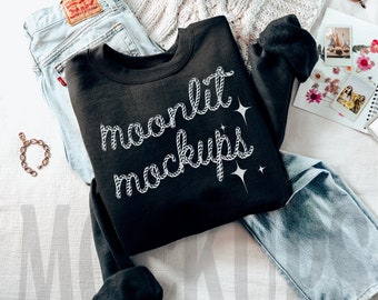 May include: A black sweatshirt with the text "moonlit mockups" in white rope-like lettering. The sweatshirt is laid out on a white knitted blanket with a pair of blue jeans and a grey backpack.