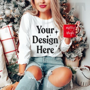 May include: A white long-sleeved shirt with the text "Your Design Here" printed on the front. The shirt is being worn by a person sitting in front of a Christmas tree and holding a red mug with the text "merry bright" printed on it.