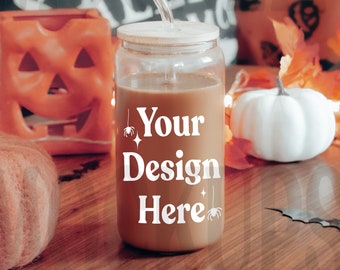 May include: A clear glass jar with a brown liquid inside and a wooden lid. The jar has a straw sticking out of the top and the text "Your Design Here" written on the side. The jar is sitting on a wooden table with a pumpkin, a white pumpkin, and a jack-o-lantern candle.