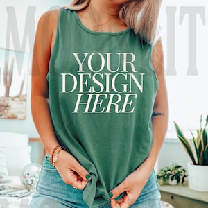 May include: A woman wearing a green tank top with the text "YOUR DESIGN HERE" printed on the front. She is wearing blue jeans and has her hands tucked into the front of the tank top.
