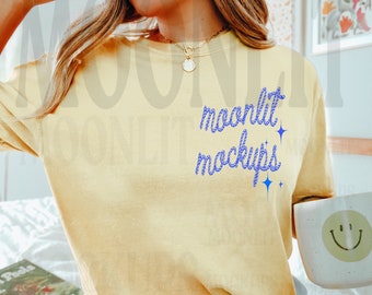May include: A pale yellow t-shirt with the words "moonlit mockups" in blue script. The person is wearing light blue shorts and holding a white mug with a smiley face. A book titled "How to Plant" is visible.