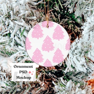 May include: A white round Christmas ornament with a pink and white pattern of stylized Christmas trees. The ornament is hanging from a gold string on a snowy artificial Christmas tree.