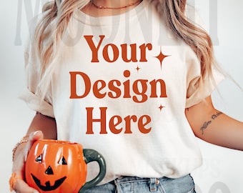 Pode incluir: Uma camiseta branca com o texto "Your Design Here" em laranja. A camiseta está sendo usada por uma pessoa que está segurando uma caneca em forma de abóbora.