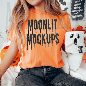 May include: A woman wearing an orange t-shirt with the text "MOONLIT MOCKUPS" in black dripping letters. She is wearing black jeans and holding a white ghost-shaped mug.