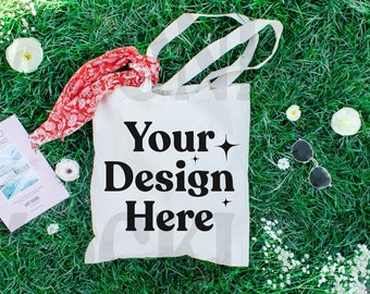May include: White canvas tote bag with black text that says "Your Design Here" on a green grassy background with white flowers and a red scarf.