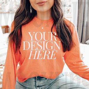 May include: A woman wearing a coral long-sleeve t-shirt with the text "YOUR DESIGN HERE" printed on the front. She is wearing blue jeans and has long brown hair.