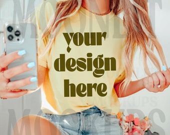 May include: A woman wearing a yellow t-shirt with the text "your design here" in brown letters. She is holding a smartphone in her left hand and her right hand is holding a strand of her blonde hair.