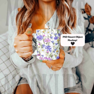 May include: White ceramic mug featuring a floral pattern with blue, purple, and white flowers. The mug has a white handle and the text "PSD Smart Object Mockup!" with a heart symbol. The mug is held in front of a person.