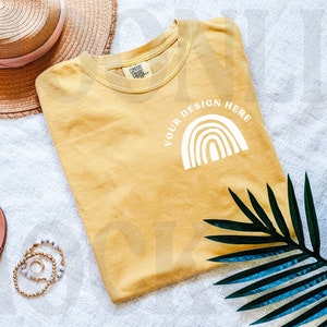 May include: A mustard yellow t-shirt with a white rainbow graphic and the text "YOUR DESIGN HERE". The t-shirt is laid flat on a white surface with a straw hat, sandals, and a green palm leaf.