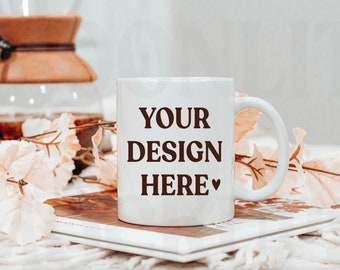 May include: White ceramic mug with brown "YOUR DESIGN HERE" text and a small heart symbol. The mug is sitting on a white surface with a book and a sprig of white flowers.