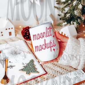 May include: White stainless steel tumbler with a red and white rope design and the text "moonlit mockups". The tumbler is being held by a hand wearing a white sweater. The tumbler is sitting on a white knitted blanket with a book and a cookie in the background.