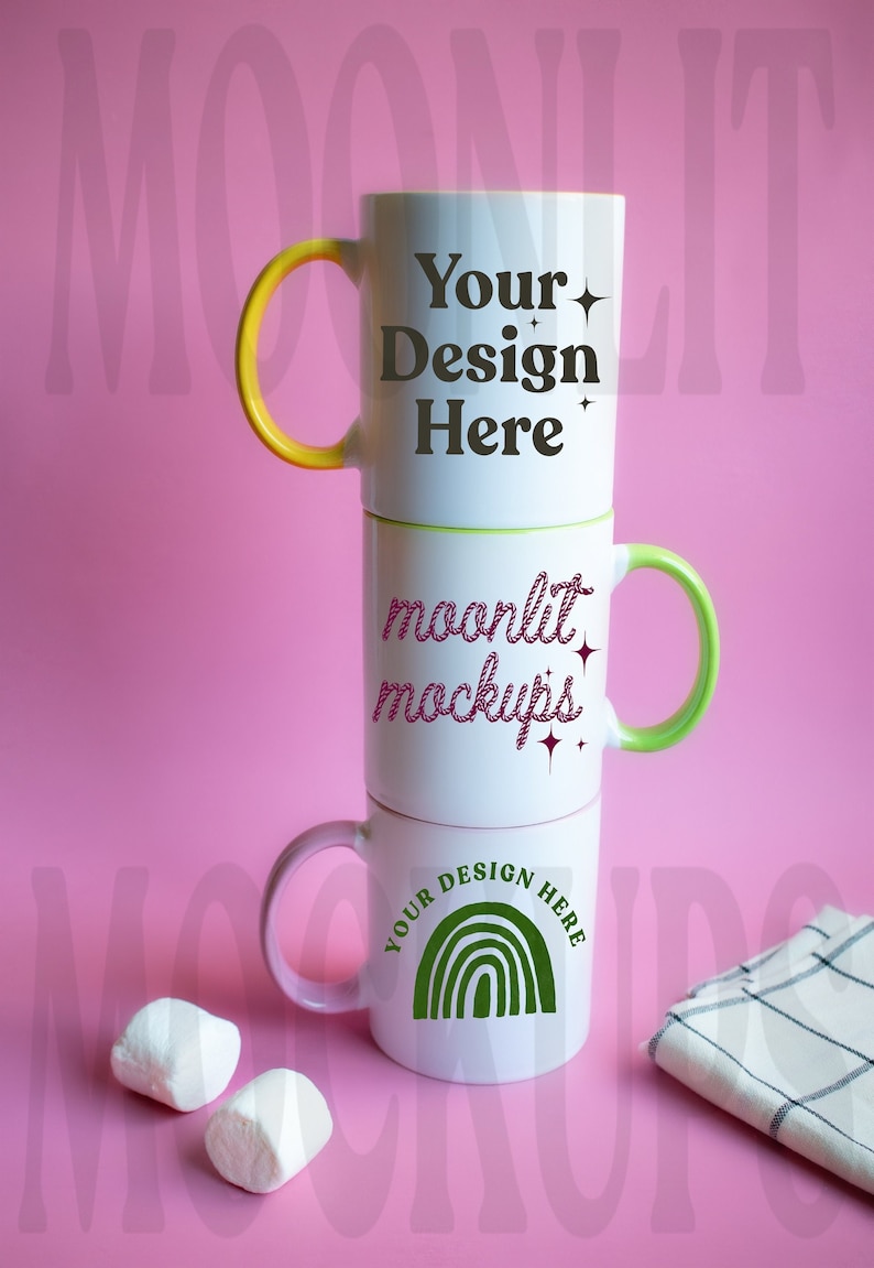 Op de afbeelding: Drie witte keramische mokken gestapeld op elkaar op een roze oppervlak. De bovenste mok heeft een gele handgreep en de tekst "Your Design Here" in zwart. De middelste mok heeft een groene handgreep en de tekst "moonlit mockups" in roze. De onderste mok heeft een roze handgreep en een groene regenboog met de tekst "Your Design Here" in zwart.