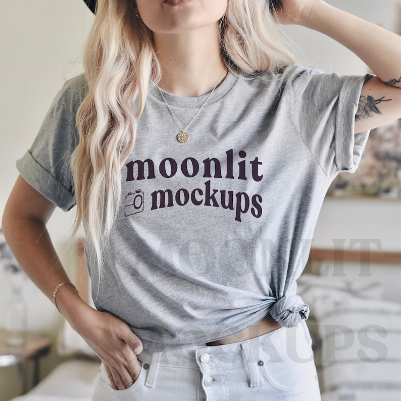 May include: A heather gray t-shirt with the text "moonlit mockups" in a dark burgundy color. The shirt has rolled-up sleeves and is knotted at the waist. The shirt is paired with white jeans.
