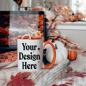 May include: White ceramic mug with an orange handle. The mug has the text "Your Design Here" in black with small stars on either side of the text. The mug is sitting on a white knitted blanket with a red and black plaid blanket underneath. There is a cinnamon stick and a slice of lemon in the mug.