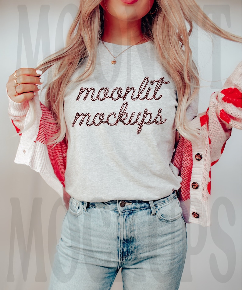 May include: A woman wearing a grey t-shirt with the text "moonlit mockups" in brown rope-like lettering. She is wearing a red and white cardigan and blue jeans.