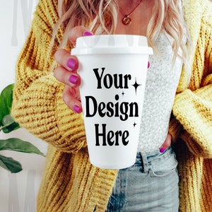 May include: A white to-go coffee cup with a black design that says "Your Design Here". The cup is being held by a person wearing a yellow cardigan and blue jeans.