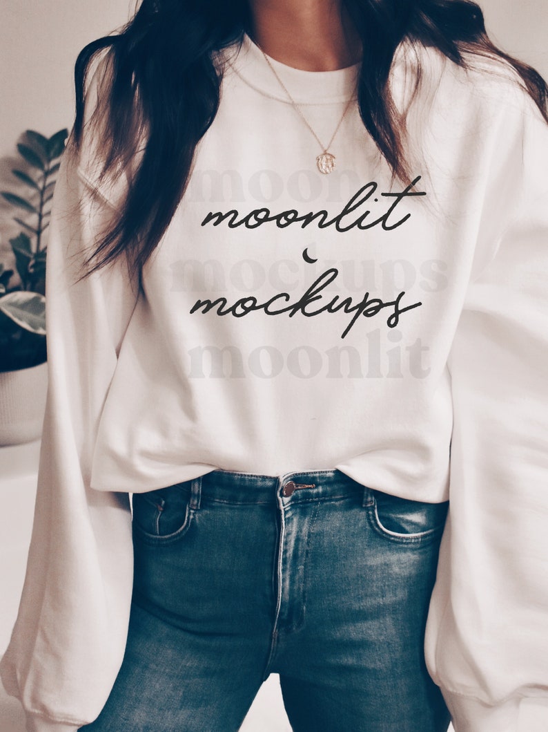 May include: A white sweatshirt with the text "moonlit mockups" in black script. The sweatshirt is worn by a person wearing blue jeans.