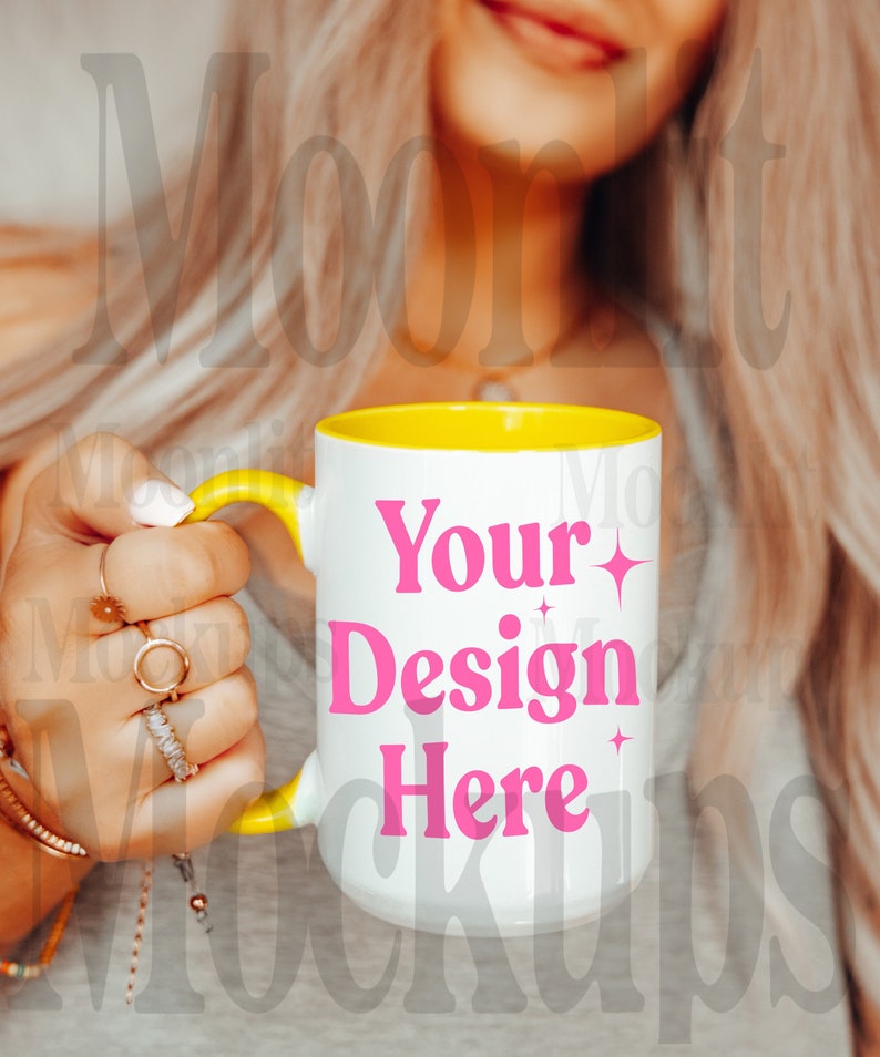 May include: White ceramic mug with a yellow interior and handle. The mug features the text "Your Design Here" in pink. The mug is held by a person with multiple rings on their fingers.