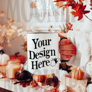 Puede incluir: Taza de cerámica blanca con borde y asa negros. La taza tiene el texto "Your Design Here" y un pequeño gráfico de calabaza impreso en ella. La taza está siendo sostenida por una mano con esmalte de uñas rojo. La taza está rodeada de decoración de otoño, incluyendo calabazas, calabazas y hojas de otoño.
