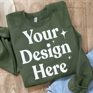 May include: A green sweatshirt with the text "Your Design Here" in white. The sweatshirt is laid flat on a wooden surface with a hat and jeans in the background.