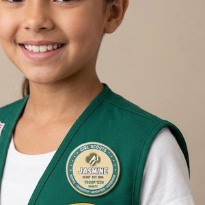 May include: A circular Girl Scouts pin featuring a cream-colored center with the name JASMINE, the text SCOUT EST. 2024, TROOP 1234, and GSNETX. The border is teal with the words GIRL SCOUTS, SERVICE, LEADERSHIP, and ADVENTURE printed in white.
