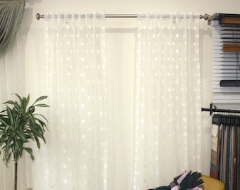 May include: Sheer white polka dot curtains hanging on a silver rod. A green and pink plaid blanket is draped over a yellow chair. A potted plant and a blue patterned rug are in the foreground.