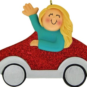 May include: A decorative ornament featuring a smiling figure with blonde hair waving from a red glitter car. The car has silver accents and black wheels. The figure is wearing a teal top.