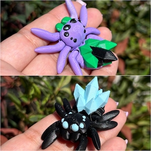 May include: Two 3D printed spider figurines. The first spider is purple with a green and black crystal on its back. The second spider is black with a light blue crystal on its back.