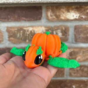 May include: A small, orange pumpkin-shaped figurine with green leaves and large black eyes. The figurine is sitting on a person's hand.