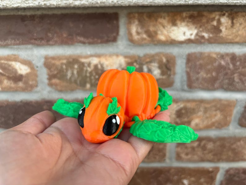 May include: A small, orange pumpkin-shaped figurine with green leaves and large black eyes. The figurine is sitting on a person's hand.