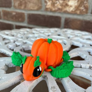 May include: A 3D printed pumpkin-shaped figurine with green leaves and black eyes. The figurine is orange and green and has a cute, cartoon-like appearance.