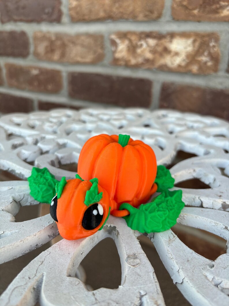 May include: A 3D printed pumpkin-shaped figurine with green leaves and black eyes. The figurine is orange and green and has a cute, cartoon-like appearance.